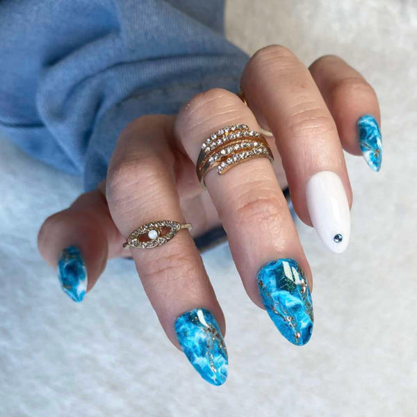 Load image into Gallery viewer, A hand wearing short almond press-on nails in a blue crystal gemstone effect design with gold accents, and delicate blue rhinestones.