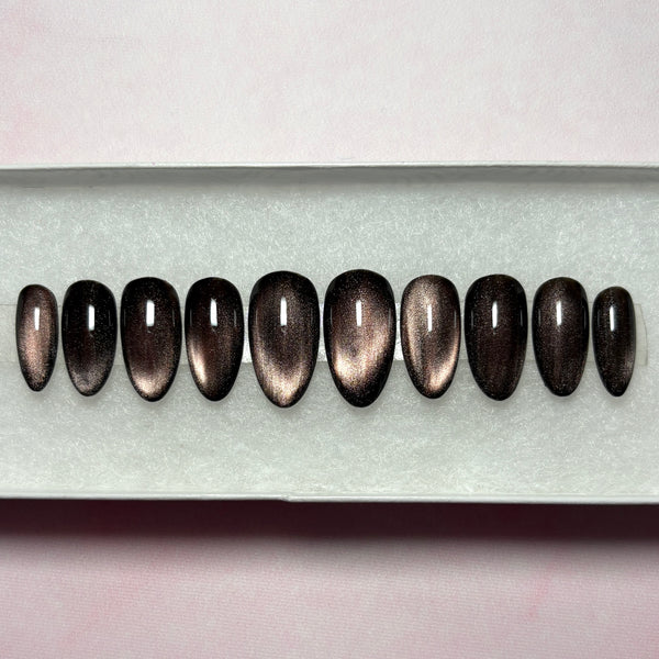 Load image into Gallery viewer, A set of false nails featuring a dark metallic cat-eye finish that shifts from burnished bronze to deep brown-black, in a short almond shape, displayed in a white presentation box.