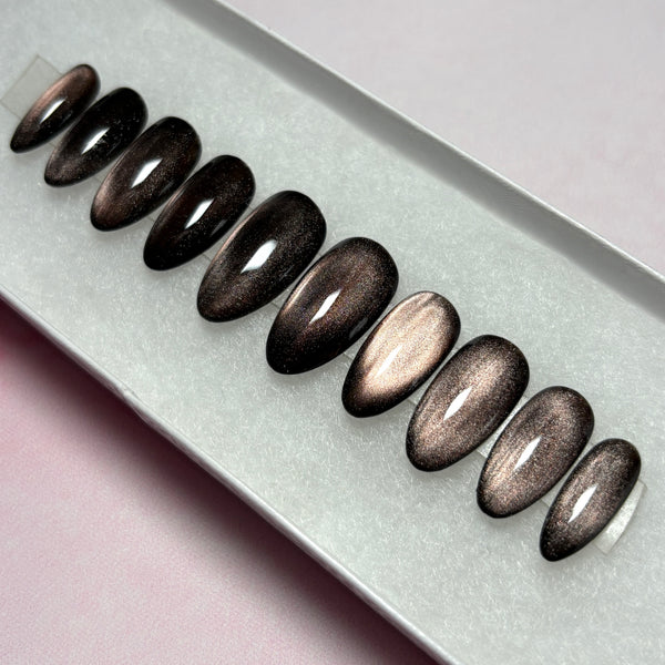 Load image into Gallery viewer, A set of stick-on nails featuring a dark metallic cat-eye finish that shifts from burnished bronze to deep brown-black, in a short almond shape, displayed in a white presentation box.