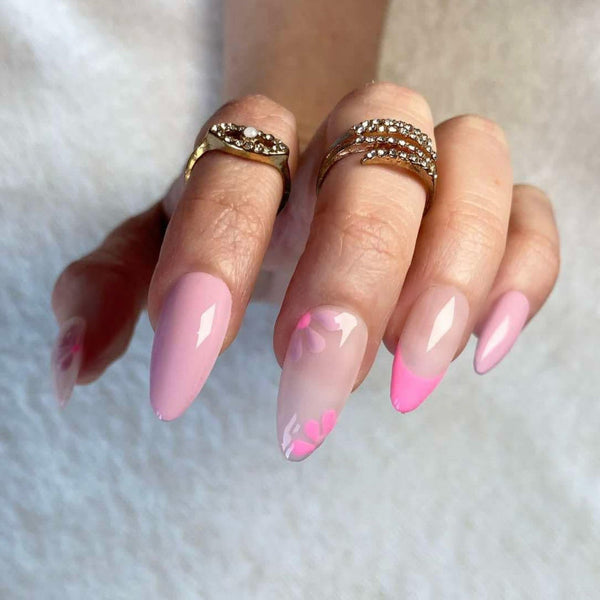 Load image into Gallery viewer, A hand showcasing long almond fake nails in varying shades of pink and lilac with floral accents and French tips, as well as two gold rings, against a white background.
