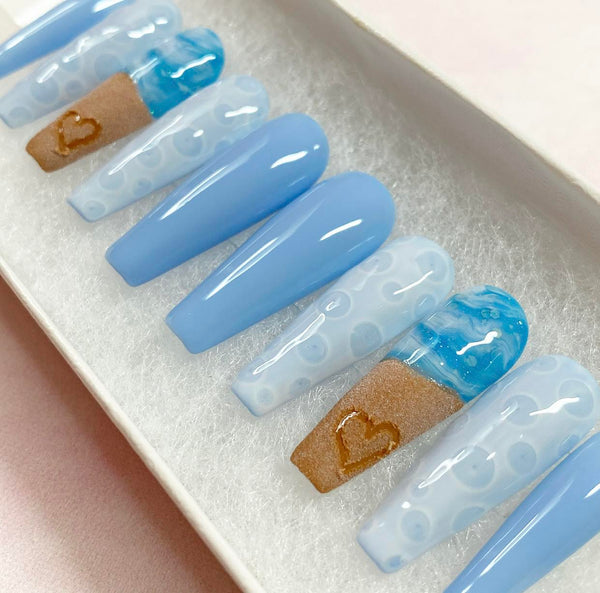 Load image into Gallery viewer, Summer press-on nails with a beach themed design featuring a realistic heart drawn in sand on two of the nails, and blue-white ocean waves and bubbles, in a long coffin shape, displayed in a white presentation box.
