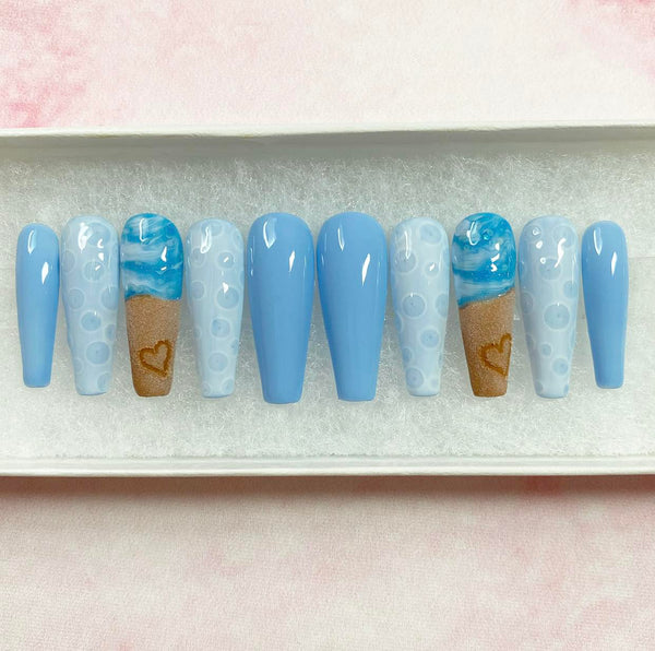 Load image into Gallery viewer, Summer holiday fake nails with a beach themed design featuring a realistic heart drawn in sand on two of the nails, and blue-white ocean waves and bubbles, in a long coffin shape, displayed in a white presentation box.