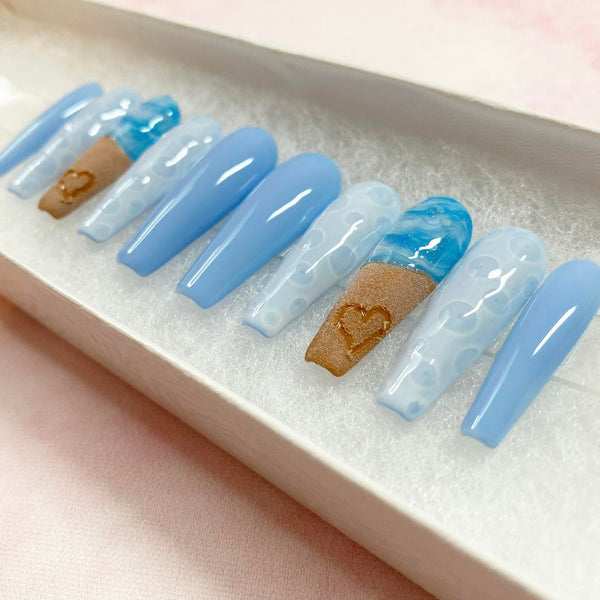 Load image into Gallery viewer, Summer press-on nails with a beach themed design featuring a realistic 3D heart drawn in the sand on two of the nails, and blue and white ocean waves and bubbles on others, in a long coffin shape.
