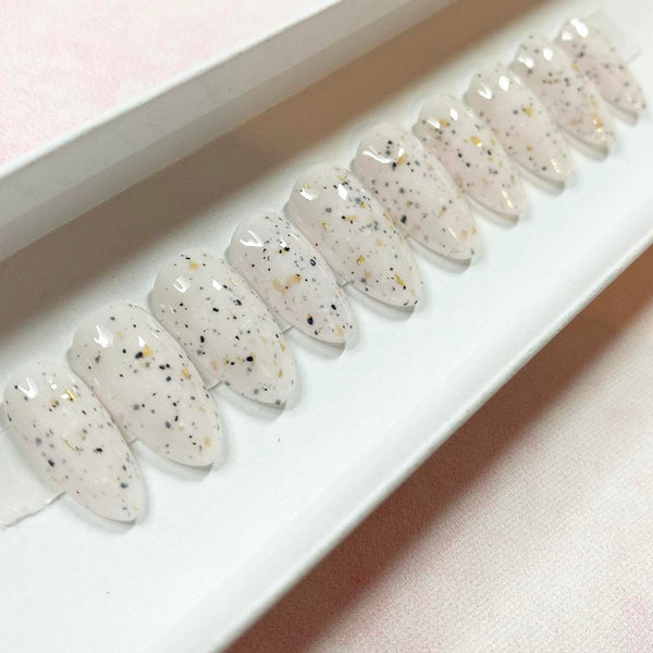 Load image into Gallery viewer, A set of jelly style press-on false nails in a pale pinkish-white base with scattered black, gray, and small gold speckles, in a short stiletto shape, displayed in a white presentation box.