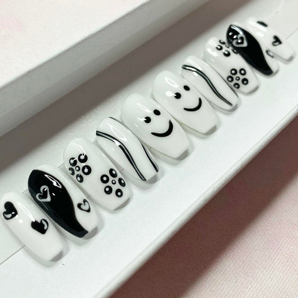 Load image into Gallery viewer, Short coffin press-on nails in black and white with various designs including tiny hearts, smiley faces and flowers, displayed in a white presentation box.