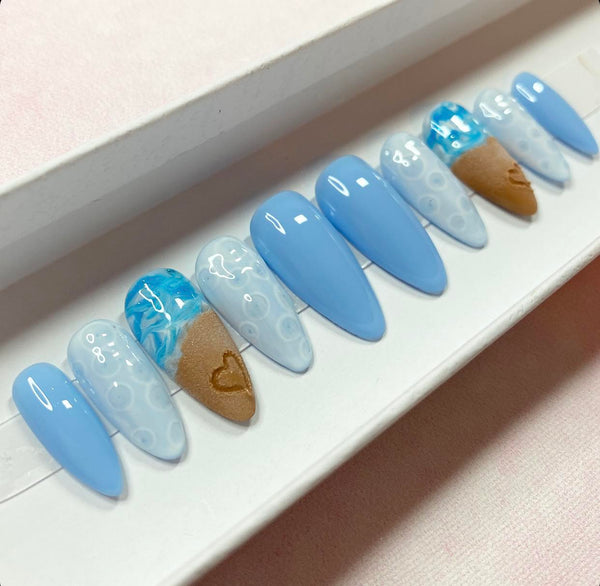 Load image into Gallery viewer, Almond shape press-on nails with a summer beach design featuring a realistic 3D heart drawn in sand on two of the nails, and blue-white ocean waves and bubbles, displayed in a white presentation box.