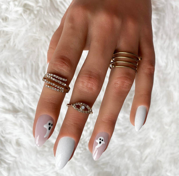 Load image into Gallery viewer, A close-up of a hand wearing Halloween press-on nails in soft pink and shimmering white with tiny white ghost designs on the pink nails. The hand is also wearing wearing multiple rings, including stacked silver rings with clear stones, a gold ring with three bands, and a gold evil eye ring with red and clear stones.