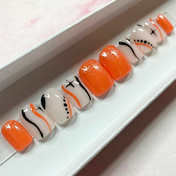 Load image into Gallery viewer, Short press-on nails with an orange and black design, featuring stars, wavy lines and dots, on a natural colour base, and subtle glitter, in a white presentation box.