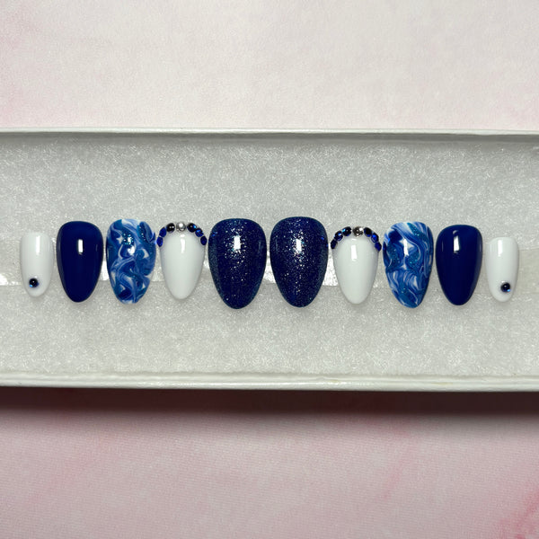 Load image into Gallery viewer, Short handmade press-on nails in navy blue and lighter blue tones, decorated with dainty blue and silver rhinestone gems, blue glitter and a marble effect, displayed in a white presentation box.