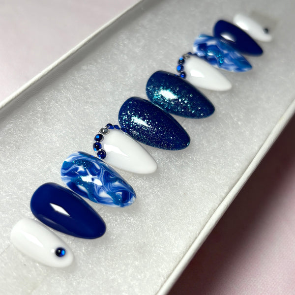 Load image into Gallery viewer, Blue press-on nails in various shades of blue and white, decorated with dainty blue and silver rhinestone gems, blue glitter and a marble effect, created by Illuminate by Liana UK.