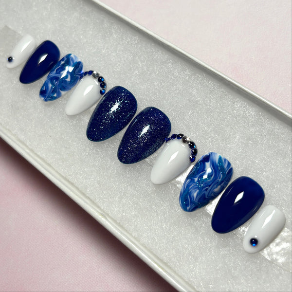 Load image into Gallery viewer, Almond shaped handmade press-on nails in navy blue and lighter blue shades, decorated with dainty blue and silver rhinestone gems, blue glitter and a marble effect, displayed in a white presentation box.