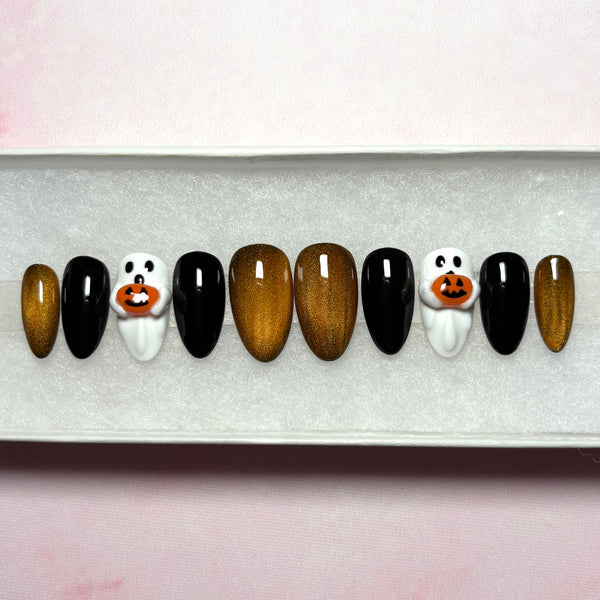 Load image into Gallery viewer, Halloween stick-on nails in a short almond shape, featuring glossy black, metallic coppery gold cat eye, and white 3D ghost designs holding tiny 3D orange jack-o'-lantern pumpkins, displayed in a white presentation box.