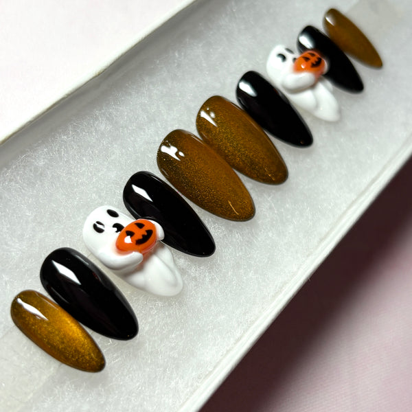 Load image into Gallery viewer, Handmade Halloween fake nails in a short almond shape, featuring glossy black, metallic coppery gold, and white 3D ghost designs holding tiny 3D orange jack-o'-lantern pumpkins, displayed in a white presentation box.