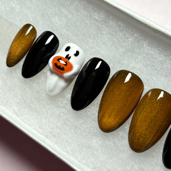 Load image into Gallery viewer, A close-up of a Halloween-themed press-on nail set displayed on a white textured tray. The set includes almond-shaped nails in a metallic gold-copper cat eye glittery finish, two glossy black nails, and a 3D white ghost nail with a cute face holding a small orange 3D jack-o'-lantern accent.