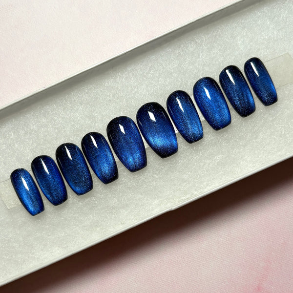 Load image into Gallery viewer, A set of blue press-on nails in cat-eye polish, that colour-shift from a shimmery dark blue through black, handmade in a short coffin shape, displayed in a white presentation box.