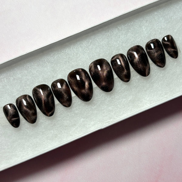 Load image into Gallery viewer, A set of ten almond-shaped press-on nails with a dark brown and black cat-eye shimmer finish, displayed in a white presentation box.