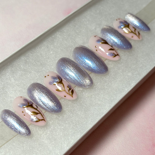 Load image into Gallery viewer, Short false nails featuring in lilac, silver and soft pink colours, with a shimmering lilac-silver cat eye, and delicate floral design with gold chrome stems and leaves, and shimmering lilac petals on a soft pink base, in a white presentation box