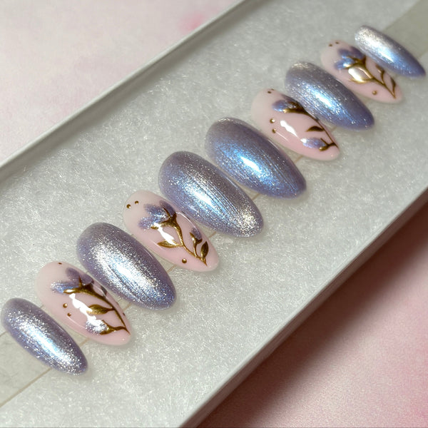 Load image into Gallery viewer, Handmade press-on nails with shimmering lilac-silver cat eye and delicate flowers with gold chrome stems and leaves, and shimmering lilac petals on a soft pink base, in a short almond shape.