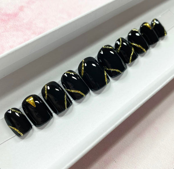 Load image into Gallery viewer, Short square handmade false nails in black, accented with gold glitter abstract lines and gold triangular-shaped gems, in a white presentation box.