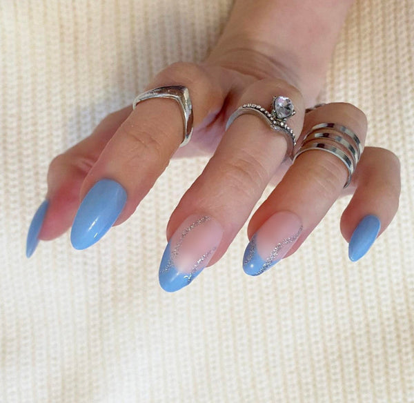 Load image into Gallery viewer, A hand wearing press-on nails featuring a baby blue colour with a French tip design and silver glitter swirl accents, in a short almond shape, as well as wearing three silver rings on the fingers, against a soft cream-coloured background.