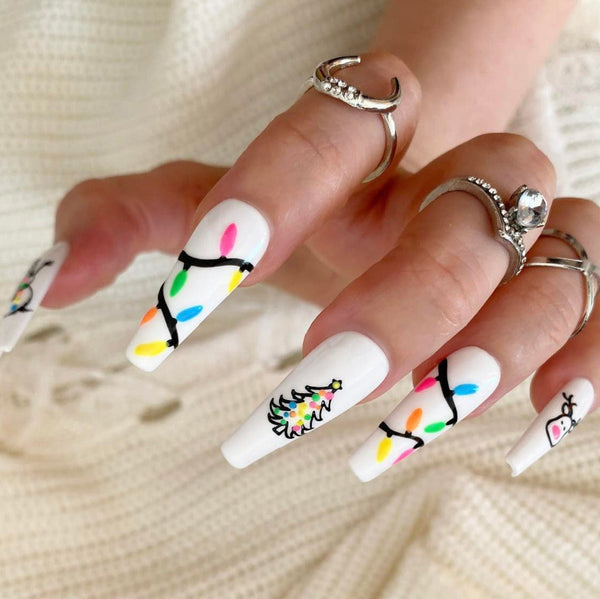 Load image into Gallery viewer, A hand wearing long coffin Christmas press-on nails that glow in the dark, featuring various Christmas designs including reindeer, fairy lights, snowmen and Christmas trees against a white background, as well as wearing three silver rings on the fingers.