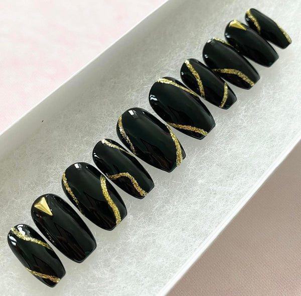 Load image into Gallery viewer, Short coffin press-on nails in black, accented with gold glitter abstract lines and gold triangular-shaped gems, in a white presentation box.