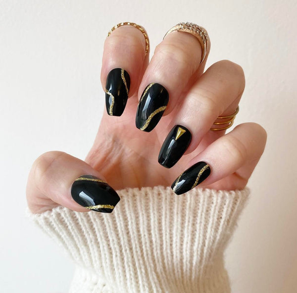 Load image into Gallery viewer, A hand showcasing press-on nails in black, accented with gold glitter abstract lines and gold triangular-shaped gems, in a short coffin shape, created by Illuminate by Liana.
