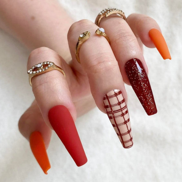 Load image into Gallery viewer, A hand wearing Autumn false nails featuring matte orange, red-orange, dark red glitter, a cream plaid with brown lines design, and a solid orange nail, in a long coffin shape, as well as wearing three gold rings on the fingers.