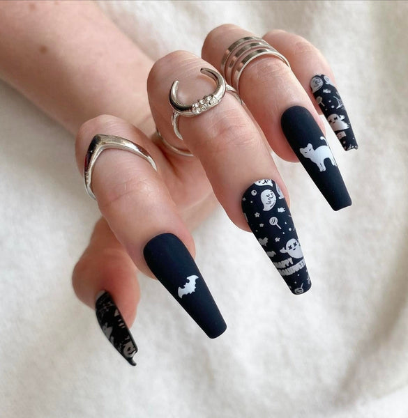 Load image into Gallery viewer, A hand wearing long coffin Halloween press-on nails featuring a black matte base with silver designs including bats, cats, ghosts, cobwebs, candy, and the wording "Happy Halloween", in a long coffin shape, as well as three rings on the fingers.
