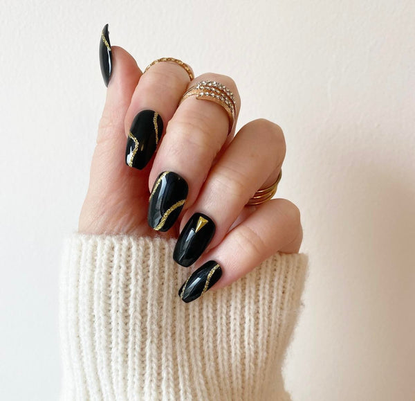 Load image into Gallery viewer, A hand wearing black press-on nails in a short coffin shape, adorned with gold glitter abstract lines and gold triangular-shaped gems, also wearing a cream coloured sleeve