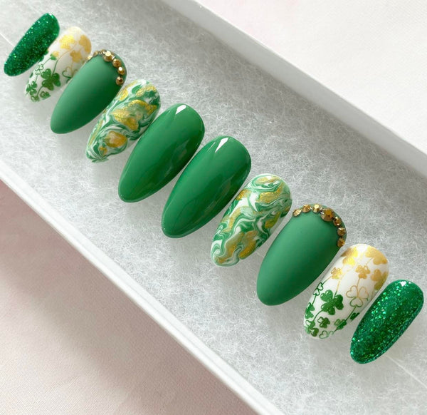 Load image into Gallery viewer, St Patrick's Day handmade short almond shaped press-on nails in green and gold featuring a green-gold shamrock design, gold rhinestones, and a gold-green marble pattern.
