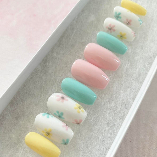 Load image into Gallery viewer, Handmade press-on nails in a short coffin shape, in pastel spring colours including yellow, blue, pink, and white, adorned with a floral design and a touch of silver glitter.