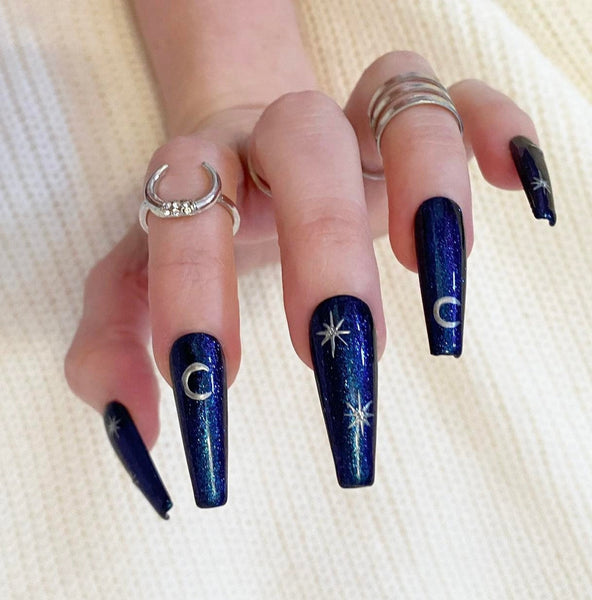 Load image into Gallery viewer, A hand wearing hHandmade false nails in deep blue with glitter and silver stars and moons artwork, giving a celestial or witchy vibe, in a long coffin shape.