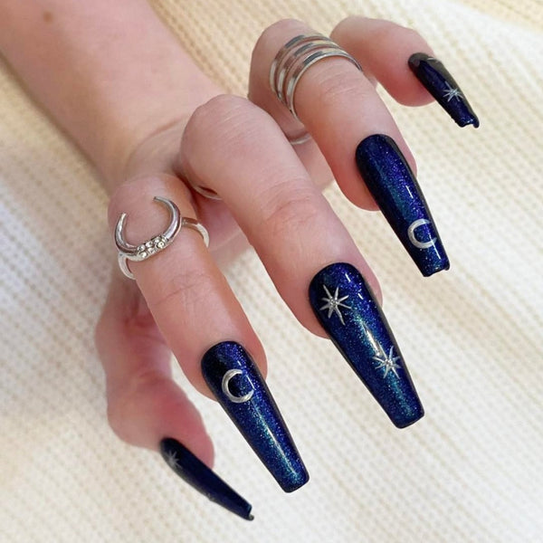 Load image into Gallery viewer, A hand wearing false nails in deep blue with glitter and silver stars and moons artwork, giving a celestial or witchy vibe, in a long coffin shape, as well as silver rings on the fingers, against a soft light background.