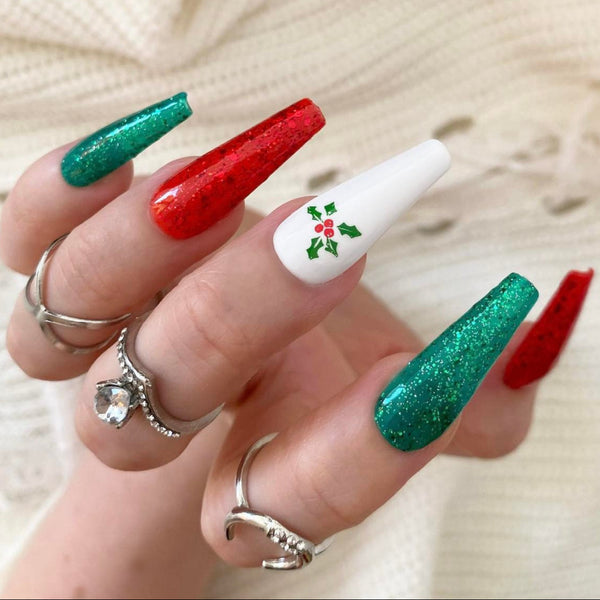 Load image into Gallery viewer, A hand wearing Christmas press-on nails in red, green and white featuring a delicate holly design, and green and red glitter, in a long coffin shape, as well as three silver rings on the fingers, against a soft cream background.