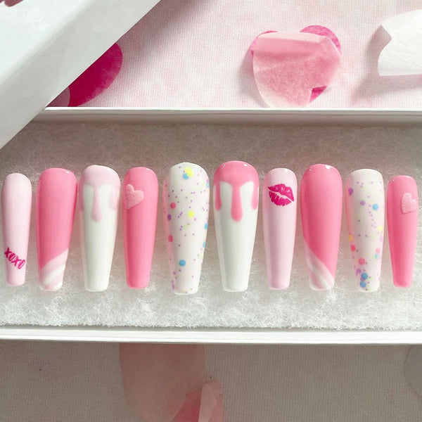Load image into Gallery viewer, Long coffin pink and white press-on nails featuring an xoxo and lips design, drip artwork, and 3D hearts all in pink, and confetti style glitter, in a white presentation box with pink petals in the background.
