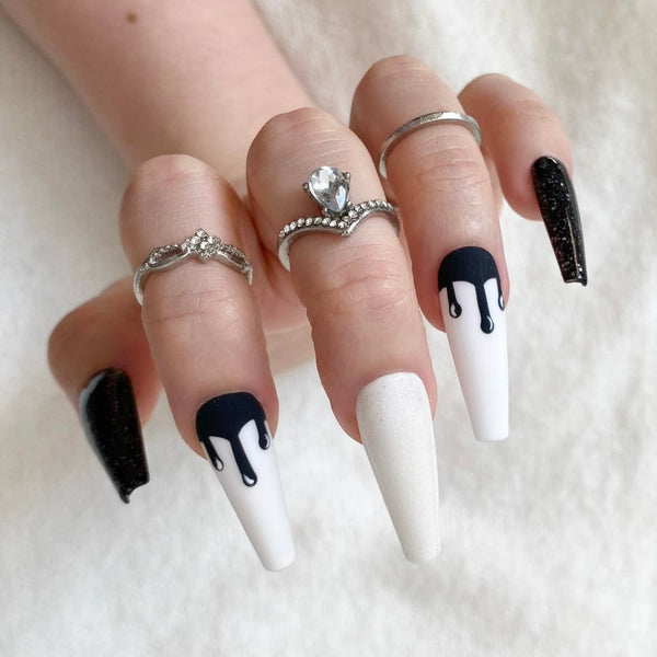 Load image into Gallery viewer, A hand displaying coffin-shaped press-on nails with a black and white drip design, pop-art style, as well as subtle white and black glitter, styled with silver rings, against a soft white background.