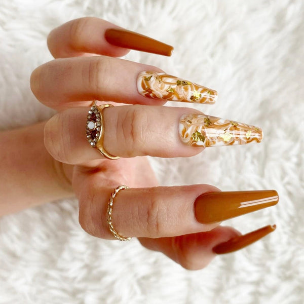 Load image into Gallery viewer, A hand wearing rings, showcasing a set of long coffin press-on nails in caramel and white with a marble design and gold foil accents.