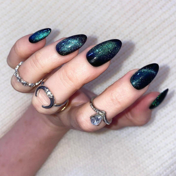 Load image into Gallery viewer, A hand showcasing press-on nails with a glittery, color-changing cat eye gel polish design in black, blue, and green shades, accompanied by various silver rings.