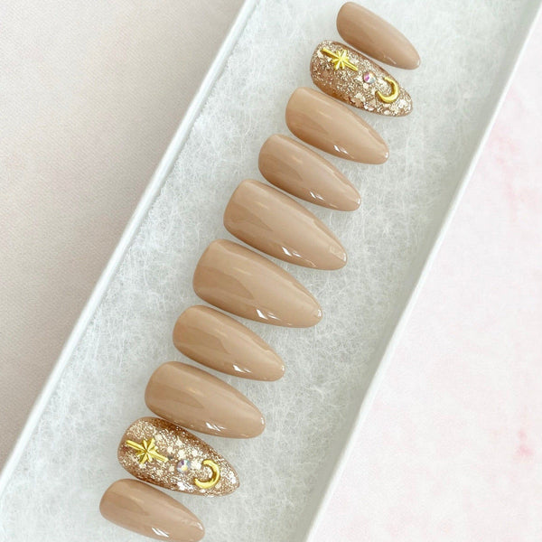 Load image into Gallery viewer, Pastel brown-nude press-on nails with two accent nails featuring gold glitter, gold crescent moon and star gems, and silver rhinestones, in a long almond shape.
