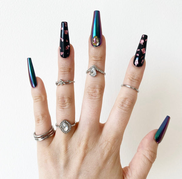 Load image into Gallery viewer, A hand wearing Halloween gothic design false nails in a long coffin shape, in a shimmering blue-green-purple holographic colour and black and pink, featuring 3D skull gems and shiny pink foil accents, as well as several silver rings on the fingers.