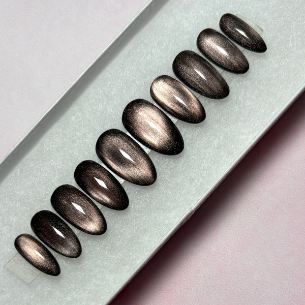 Load image into Gallery viewer, Handmade press-on nails featuring a dark metallic cat-eye finish that shifts from burnished bronze to deep brown-black, in a short almond shape, displayed in a white presentation box.
