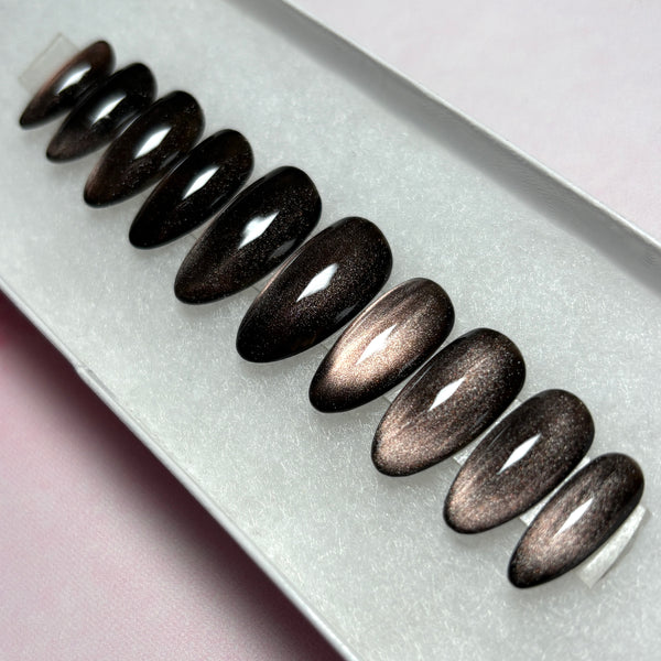 Load image into Gallery viewer, Cat eye false nails featuring a dark metallic finish that shifts from burnished bronze to deep brown-black, in a short almond shape, displayed in a white presentation box.
