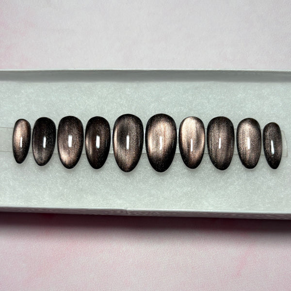 Load image into Gallery viewer, Handmade stick-on nails featuring a dark metallic cat-eye finish that shifts from burnished bronze to deep brown-black, in a short almond shape, displayed in a white presentation box.
