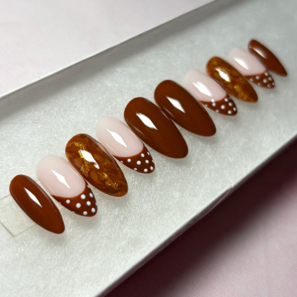 Load image into Gallery viewer, French tip stick-on nails in a short almond shape, in warm chocolate brown, featuring four solid brown nails, four French tip nails with chocolate-brown tips adorned with white polka dots, and two marble effect brown and metallic gold nails, displayed in a white presentation box.

