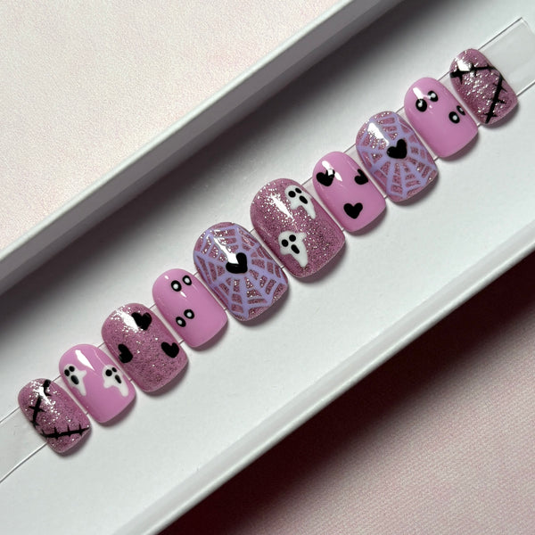 Load image into Gallery viewer, A set of Halloween-themed press-on nails displayed in a white presentation box. The nails feature a light pink and lavender color palette with spooky designs including spiderwebs, ghosts, hearts, and scars, with some nails featuring shimmering pink and silver glitter.
