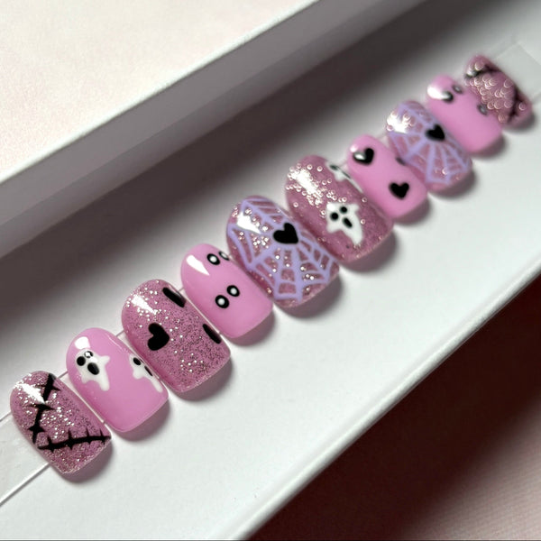 Load image into Gallery viewer, Halloween press-on nails in a short square shape, featuring pink glitter, lavender spiderwebs with black hearts at their centres, white ghosts, and black hearts and scars, displayed in a white presentation box.
