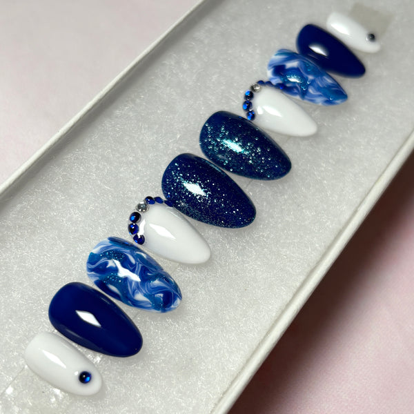 Load image into Gallery viewer, Short almond press-on nails in various shades of blue and white, featuring dainty blue and silver rhinestone gems, blue glitter and a marble effect.
