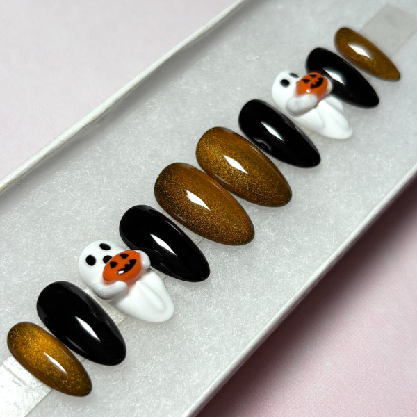 Load image into Gallery viewer, Halloween handmade press-on nails in a short almond shape, featuring glossy black, metallic coppery gold cat eye polish, and white 3D ghost designs holding tiny 3D orange jack-o'-lantern pumpkins, displayed in a white presentation box.
