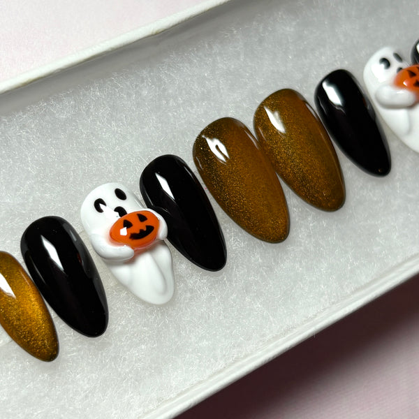 Load image into Gallery viewer, A close-up of a set of Halloween press-on nails in a short almond shape, featuring glossy black, metallic coppery gold, and white 3D ghost designs holding tiny 3D orange jack-o'-lantern pumpkins, displayed in a white presentation box.
